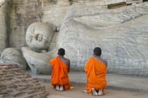 Dois monges budistas orando na frente de uma estatua de um Buda adormecido.