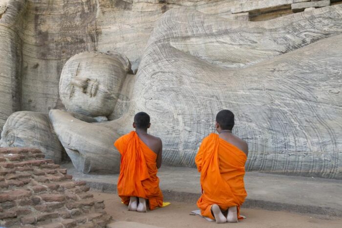 Dois monges budistas orando na frente de uma estatua de um Buda adormecido.